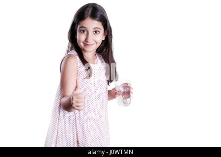 Bella felice bambina con lunghi capelli scuri e il vestito tenendo un bicchiere di acqua e mostrando il pollice. studio shot, isolato su sfondo bianco. Foto Stock