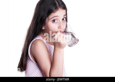 Bella felice bambina con lunghi capelli scuri e abiti azienda bicchiere d'acqua. L'acqua potabile e guardando la fotocamera. studio shot, isolato su bianco Foto Stock