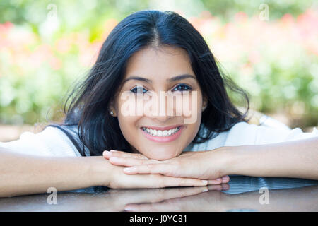 Closeup ritratto sorride fiduciosa piuttosto giovane donna faccia di appoggio sulle mani in una camicia bianca, isolato sfondo fiori sfocata. Positivo emotio umana Foto Stock