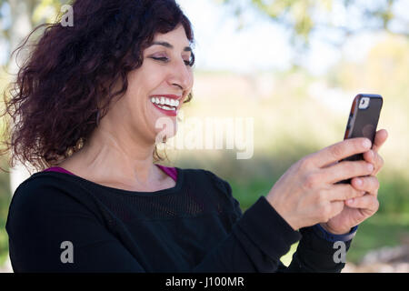 Closeup ritratto, giovani happy business donna in maglia nera in piedi, controllare il suo cellulare, isolato su sfondo sfondo all'aperto. business com Foto Stock