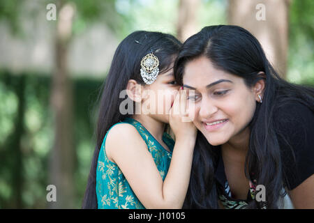 Closeup ritratto, figlia sussurra segreti in mom l orecchio, isolata all'esterno al di fuori di verdi alberi sullo sfondo Foto Stock