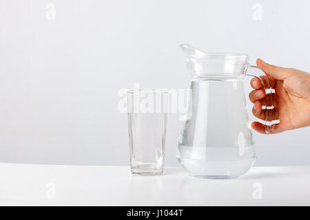 Mano versando acqua dalla caraffa di vetro di vetro contro uno sfondo bianco Foto Stock