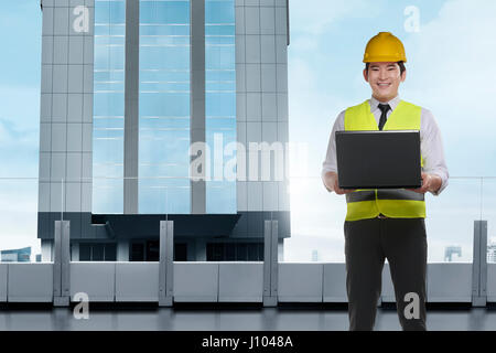 Bello architetto asiatico Uomo con notebook indossare casco protettivo in piedi di fronte ad un edificio di grattacieli Foto Stock