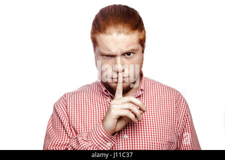 Imprenditore serio con il dito sulle labbra facendo silenzio gesto. shhh! Readhead barbuto imprenditore con maglietta rossa e lentiggini guardando la telecamera prigioniero. Foto Stock