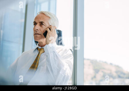 Senior business uomo seduto alla sua scrivania e parlando al telefono cellulare. Senior manager utilizzando il cellulare in ufficio. Foto Stock