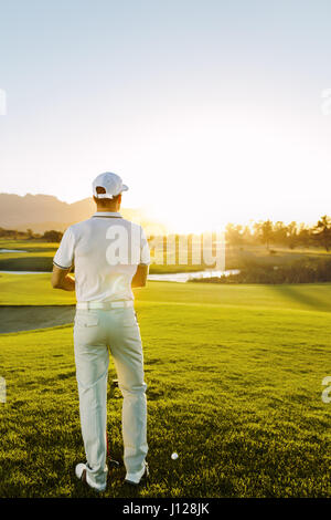 Vista posteriore di un giovane maschio golfista permanente al quadrato con un raccordo a T sul corso. Professional Golfer maschio giocando in una giornata di sole. Foto Stock