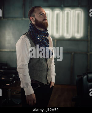 Bel uomo barbuto hipster con barba elegante in una camicia bianca e chekered sciarpa in piedi con la faccia di gravi nel salone di bellezza o di Barberia Foto Stock