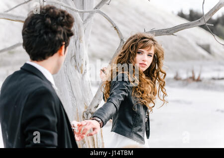 Giovane donna con capelli ricci tenendo la mano del giovane uomo in piedi torna alla telecamera vicino all'Albero appassito nel deserto. Foto Stock