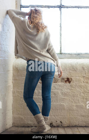 Slim donna che indossa jeans in piedi dalla finestra appoggiata contro il muro Foto Stock