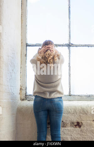 Vista posteriore di un sottolineato donna bionda in piedi dalla finestra in testa le mani Foto Stock