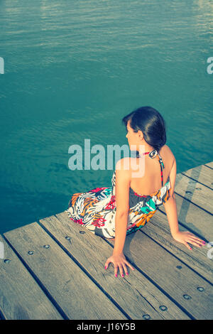 Vista posteriore di una giovane donna si sedette sul pontile sul mare Foto Stock