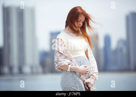 Redheaded donna incinta toccando il suo ventre con grattacieli in background. Estate Foto Stock