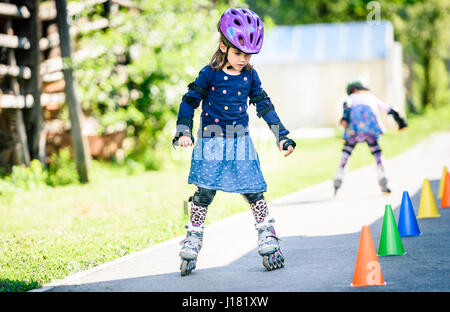 I bambini che imparano a pattino a rotelle sulla strada con i coni. Due gemelle sono la pratica di sicuro pattinaggio su una casa carraio strada indossando gea protettivo Foto Stock