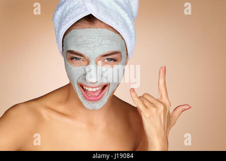 Funny donna che indossa una maschera per il viso e fare rock gesto a mano. Ritratto di giovane donna su sfondo beige. Cura della Pelle concept Foto Stock