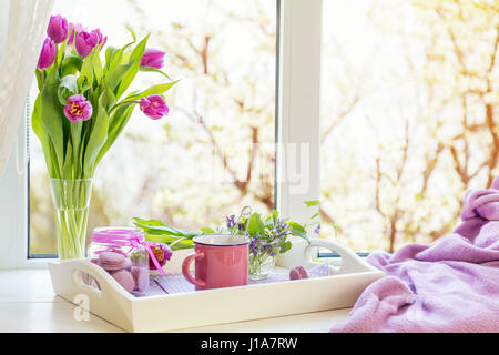 Accogliente casa concetto. Viola tulipani freschi in vaso di vetro. Amaretti in un barattolo di vetro. Tazza di tè caldo. Vassoio bianco Coperta di Lilla sul davanzale. Sunshine. C Foto Stock