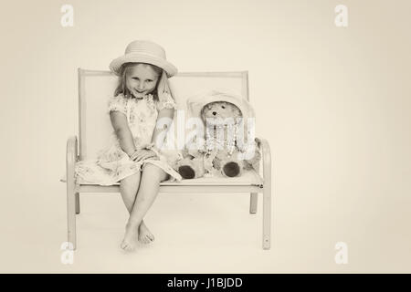 Bambina il bambino che indossa un floral abiti estivi e cappello di paglia con abbinamento di Teddy seduto su una biposto panchina Foto Stock