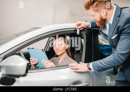 Barbuto venditore dando chiave auto per la giovane donna seduta nella nuova vettura con tavoletta digitale Foto Stock