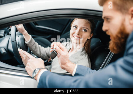 Barbuto venditore dando chiave auto a sorridente giovane donna seduta nella nuova vettura Foto Stock