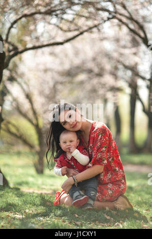 Giovane madre con il suo bambino sulla passeggiata nel giardino di primavera. Essi sono qui seduti su erba tra gli alberi. Foto Stock