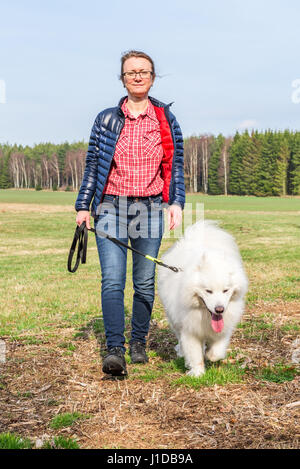 Gestore femmina e il suo cane Samoiedo camminando verso di te da un campo nella foresta. Foto Stock