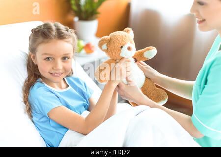 Ritagliato shot di infermiere dando orsacchiotto per sorridente bambina in ospedale Foto Stock