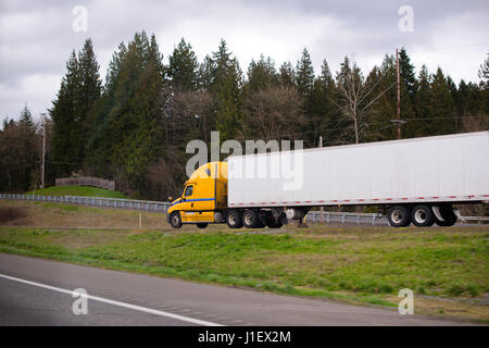 Modern big rig giallo luminoso semi carrello per i voli di lungo raggio con lunghi dry van rimorchio per il trasporto di carichi secchi prendere l'uscita per la collina sulla autostrada sempreverdi. Foto Stock