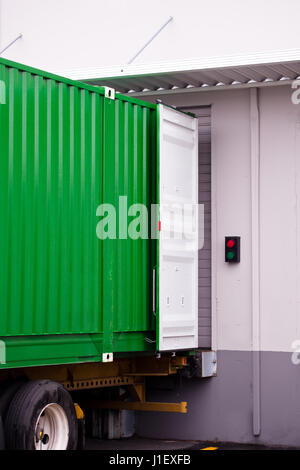 Un grande contenitore di metallo verde all'esterno con il bianco porta aperta su un semi camion rimorchio in giallo in un dock stazionaria magazzino con una tettoia in metallo Foto Stock