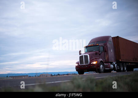 Maroon classica big rig semi carrello con fari anteriori trasporta il contenitore sulla strada che corre lungo gli edifici industriali e commerciali in sera Foto Stock