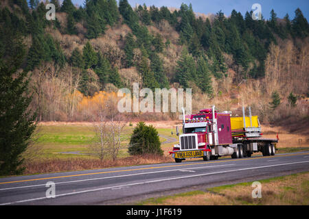 Americano classico rosso semi carrello con una griglia di grande e alto di statura tubazioni di scarico e di una cabina lunga e un segno over size trasporti di carico lungo una strada panoramica cargo Foto Stock