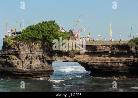 Naturale stupendo arco di roccia a pura Batu Bolong in Tanah Lot area del tempio con i pellegrini in abito cerimoniale passando attraverso il foro nella roccia a Bali Foto Stock