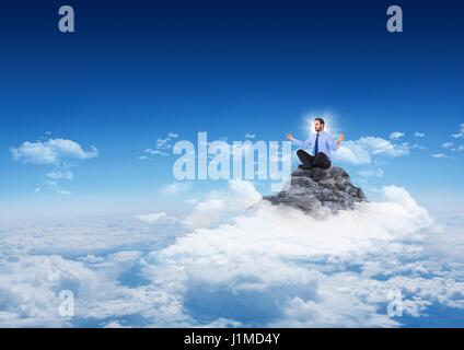 Composito Digitale dell uomo d affari con la torciera meditando sul picco di montagna tra le nuvole con cielo blu Foto Stock