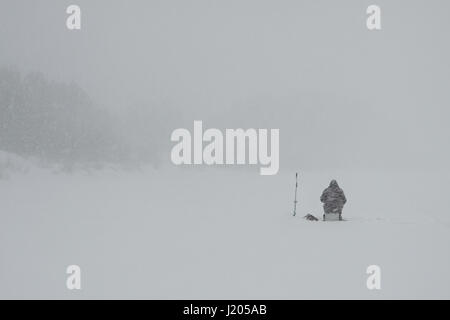 Una vista del pescatore solitario seduto sul fiume icebound e pesca durante una nevicata Foto Stock