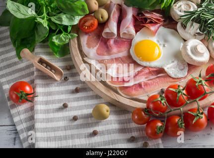 Una sana colazione mediterranea alimentari: uova fritte, prosciutto, pomodori a grappolo, funghi, basilico, olive, rosmarino sul vassoio in legno Foto Stock