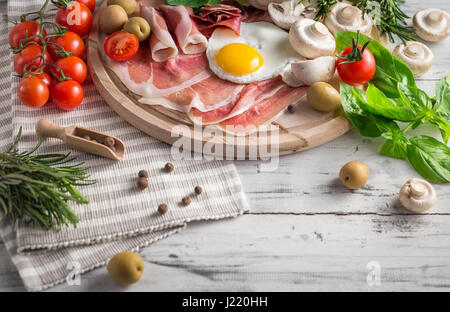 Una sana colazione mediterranea alimentari: uova fritte, prosciutto, pomodori a grappolo, funghi, basilico, olive, rosmarino sul vassoio in legno Foto Stock