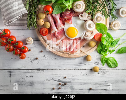 Una sana colazione mediterranea alimentari: uova fritte, prosciutto, pomodori a grappolo, funghi, basilico, olive, rosmarino sul vassoio in legno Vista da sopra Foto Stock