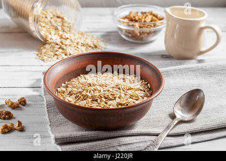 Fiocchi di avena in argilla marrone ciotola pronto per cucinare, sparsi in fiocchi di avena, le noci, la caraffa con il latte e versare sul light tavolo in legno Foto Stock