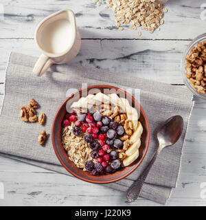 Farina di avena in argilla marrone ciotola con frutti di bosco surgelati (ribes nero, ribes, more), banane e noci pronto a mangiare, la caraffa con il latte e il cucchiaio o Foto Stock