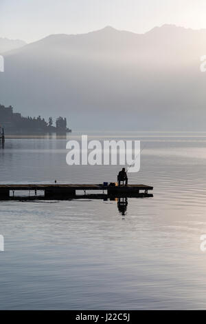 Pescatore solitario seduto sul molo pesca su un inizio di foschia mattutina a Lenno, Lago di Como, Italia nel mese di aprile Foto Stock