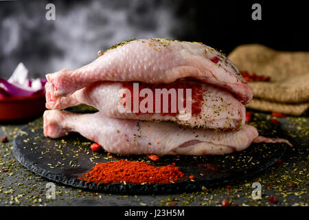 Primo piano di alcune materie le cosce di pollo su una piastra di ardesia, posto su una tavola in legno rustico cosparso con diverse spezie Foto Stock