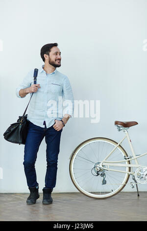 Ritratto di contemporary barbuto uomo asiatico che guarda lontano e sorridente felicemente in piedi contro la parete con la bici vestito in abiti casual Foto Stock