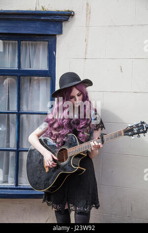 Un giovane suonatore ambulante femmina con lunghi capelli viola,cantando e suonando la chitarra a Whitby Goth Weekend in North Yorkshire, Inghilterra, Regno Unito Foto Stock