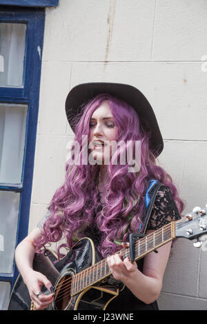 Un giovane suonatore ambulante femmina con lunghi capelli viola,cantando e suonando la chitarra a Whitby Goth Weekend in North Yorkshire, Inghilterra, Regno Unito Foto Stock