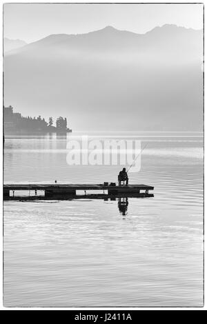 Pescatore solitario seduto sul molo pesca su un inizio di foschia mattutina a Lenno, Lago di Como, Italia nel mese di aprile Foto Stock