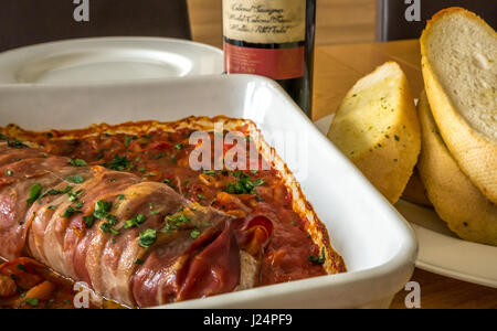 Close up italiane di filetti di maiale su letto di pomodori, fagioli e aglio con la piastra di aglio pane, un bicchiere di vino rosso, e la bottiglia di vino sul tavolo di quercia Foto Stock