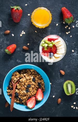 Muesli con noci e uvetta, bicchiere di yogurt con frutta fresca, un vasetto di miele e più fresche Fragole e kiwi in pietra scura sullo sfondo. In verticale Foto Stock