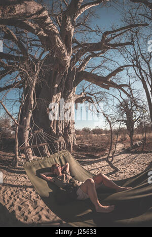 Giovane tenendo selfie sull amaca appesa da enormi Baobab nella savana africana. Vista fisheye, tonica immagine. Wilderness Safari e avventura in un Foto Stock