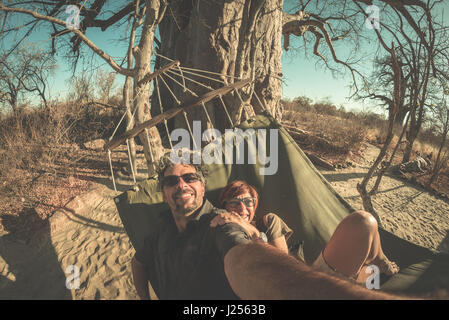 Giovane tenendo selfie sull amaca appesa da enormi Baobab nella savana africana. Vista fisheye, tonica immagine. Wilderness Safari e avventura in un Foto Stock