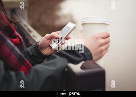 Immagine ritagliata della donna utilizzando il telefono cellulare mentre si tiene tazza monouso Foto Stock