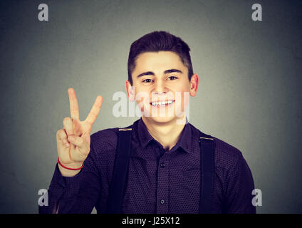 Ritratto felice ragazzo adolescente che mostra la vittoria o segno di pace isolato sul muro grigio Sfondo. Faccia positiva espressione emozione il linguaggio del corpo Foto Stock