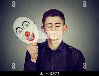 Giovane uomo triste decollare happy clown maschera isolata sul muro grigio Sfondo. Le emozioni umane. Personalità scissa concept Foto Stock